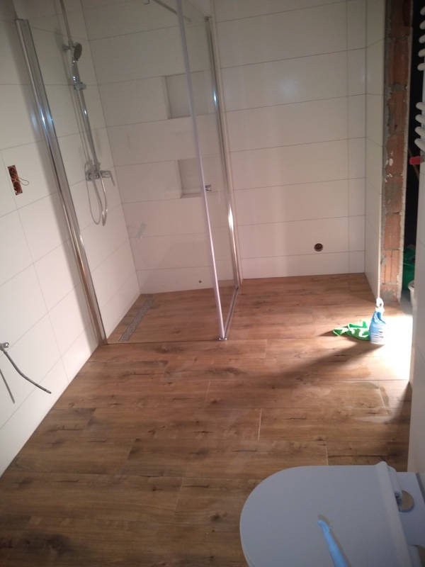 Wnętrze łazienki w trakcie prac wykończeniowych: białe płytki na ścianach, drewnopodobna podłoga, częściowo zamontowana kabina prysznicowa z chromowanymi profilami, widoczne niedokończone...