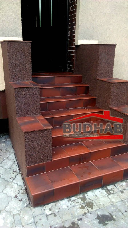 Wykończone płytkami schody wejściowe z brązowymi płytkami i kolumnami o chropowatej fakturze, widoczne logo firmy BUDHAB na schodach.