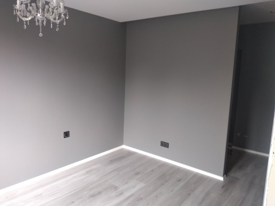 Pokój po malowaniu na szaro z drewnopodobną podłogą, widoczny żyrandol kryształowy i czarne gniazdka elektryczne.