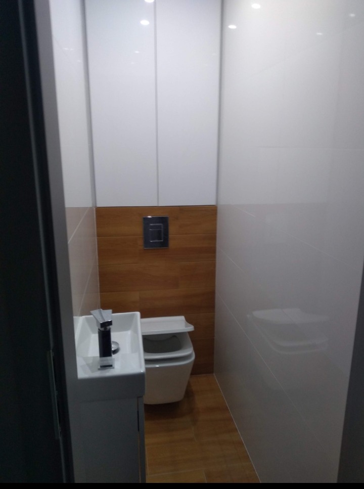 Wąska toaleta z umywalką na szafce, drewnopodobne panele na ścianie do połowy wysokości, białe płytki i szafki wiszące, minimalistyczny design.