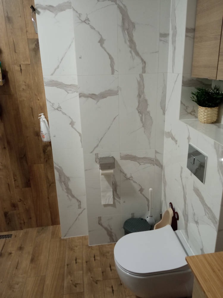 Nowoczesna łazienka z białymi płytkami imitującymi marmur, drewnianą podłogą i toaletą. Widoczny pojemnik na papier toaletowy oraz szczotka do WC w rogu.