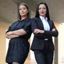 Dwie kobiety w eleganckich strojach stojące plecami do siebie z założonymi rękami, prezentujące pewność siebie i profesjonalizm.