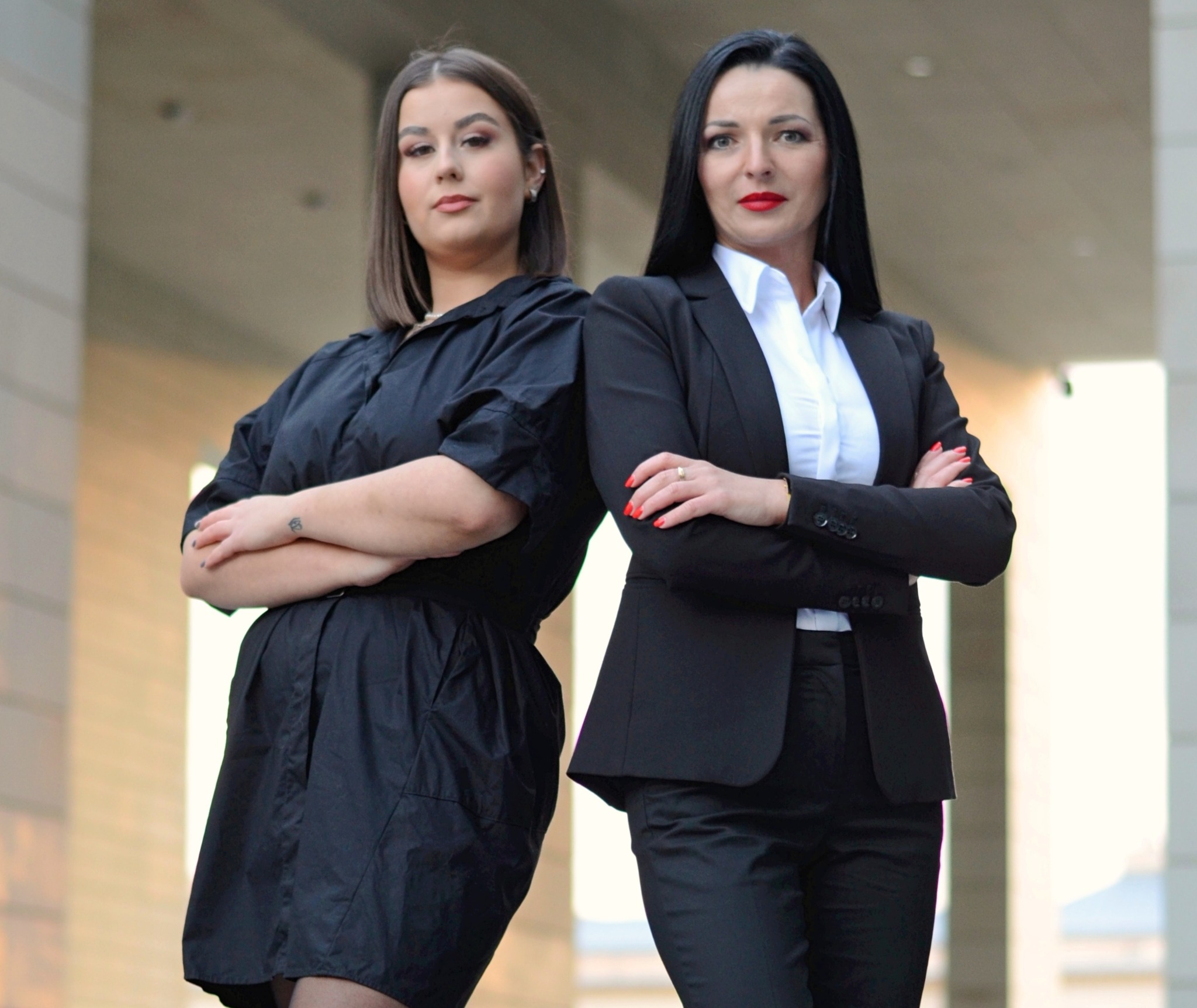 Dwie kobiety w eleganckich strojach stojące plecami do siebie z założonymi rękami, prezentujące pewność siebie i profesjonalizm.