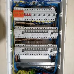 Tablica rozdzielcza z zamontowanymi bezpiecznikami i okablowaniem w kolorach niebieskim, żółtym i zielonym, widoczna w białej obudowie w ścianie.