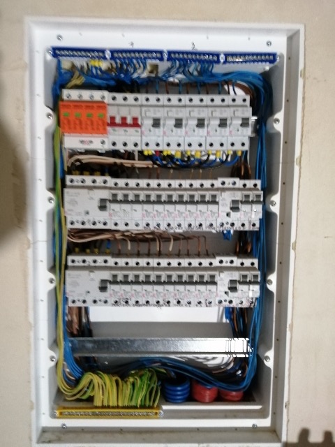 Tablica rozdzielcza z zamontowanymi bezpiecznikami i okablowaniem w kolorach niebieskim, żółtym i zielonym, widoczna w białej obudowie w ścianie.
