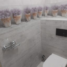 dolecki dariusz - Wykończona łazienka z szarymi płytkami imitującymi drewno, dekoracyjnym paskiem z motywem lawendy w doniczkach, wiszącą toaletą i chromowanym uchwytem na papier.