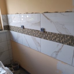dolecki dariusz - Fragment ściany w trakcie kładzenia płytek w kuchni lub łazience, widoczne białe płytki imitujące marmur i dekoracyjny pas mozaiki, puszki elektryczne wystające ze ściany, ślady kleju...