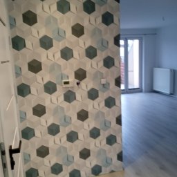 dolecki dariusz - Ściana wykończona dekoracyjną tapetą w geometryczne wzory, widoczny fragment drzwi z klamką oraz fragment podłogi z panelami, w tle jasne pomieszczenie z oknem i grzejnikiem.