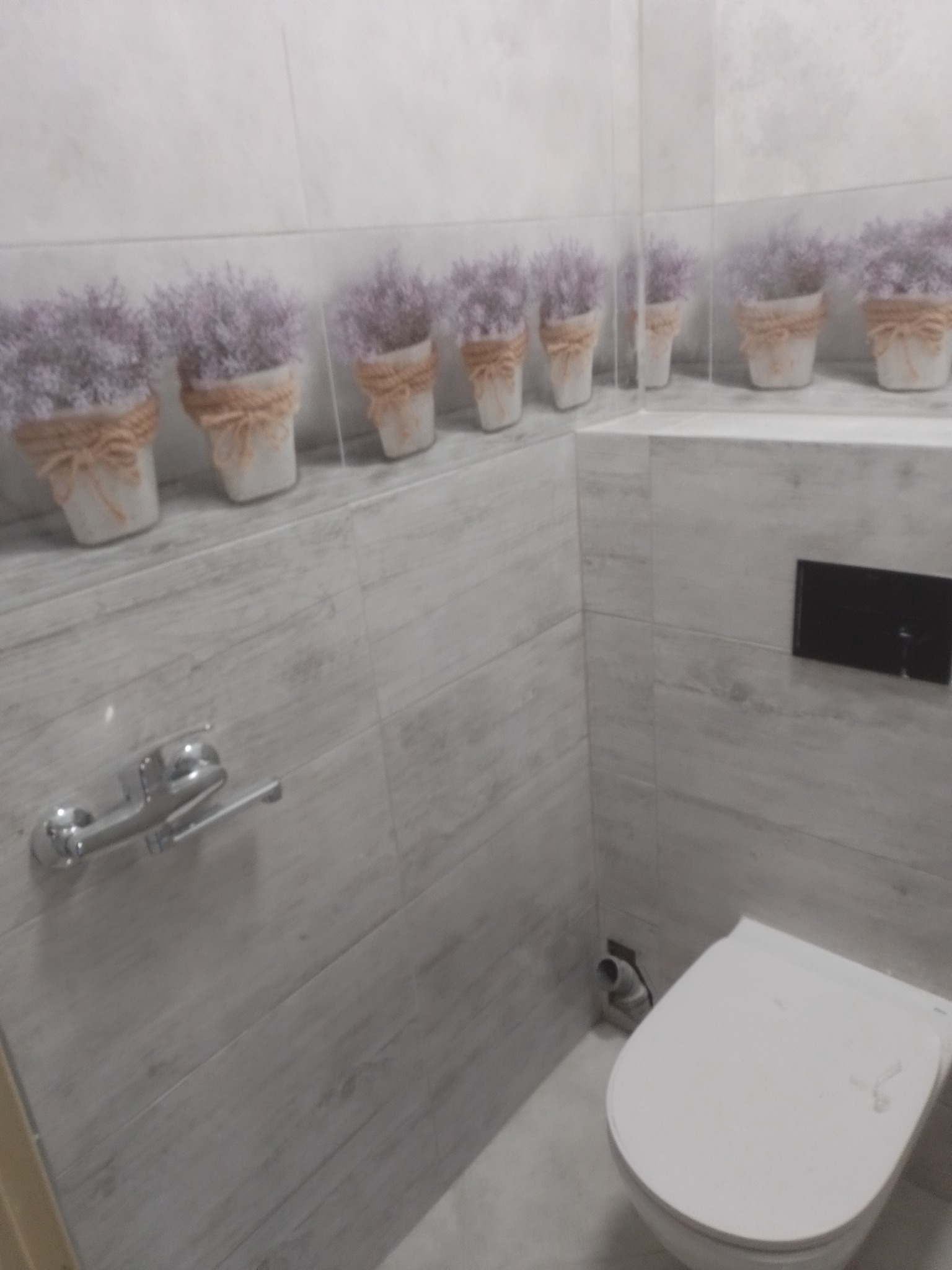 Wykończona łazienka z szarymi płytkami imitującymi drewno, dekoracyjnym paskiem z motywem lawendy w doniczkach, wiszącą toaletą i chromowanym uchwytem na papier.