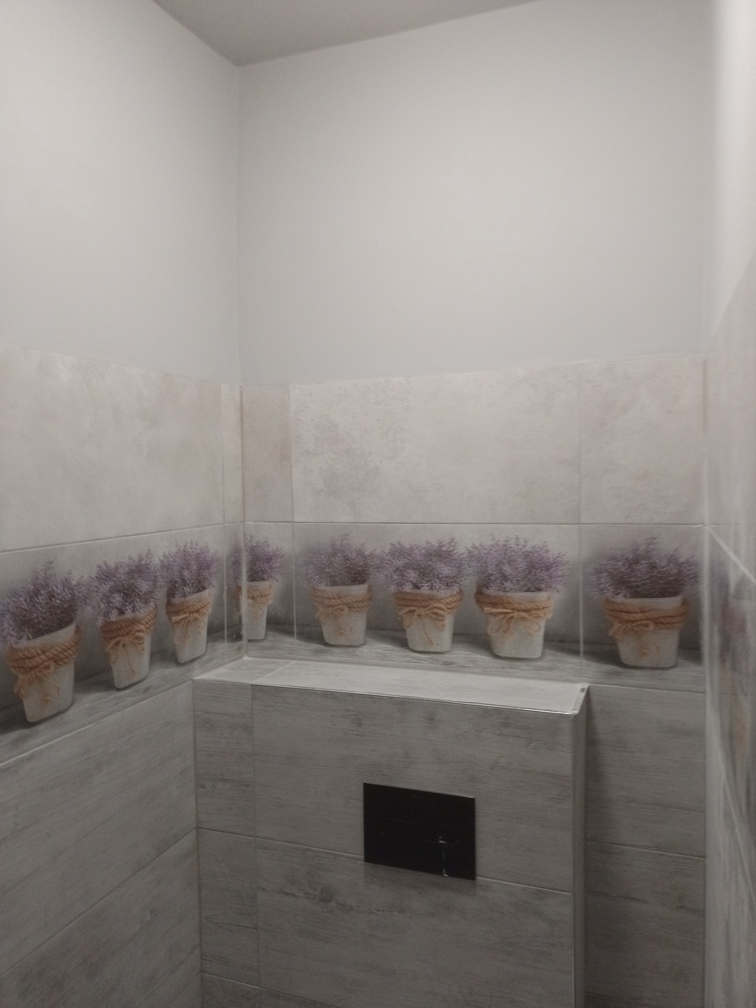 Wykończona łazienka z szarymi płytkami imitującymi drewno i dekoracyjnym motywem lawendy w doniczkach, umieszczonym wzdłuż ściany nad zabudową toalety.