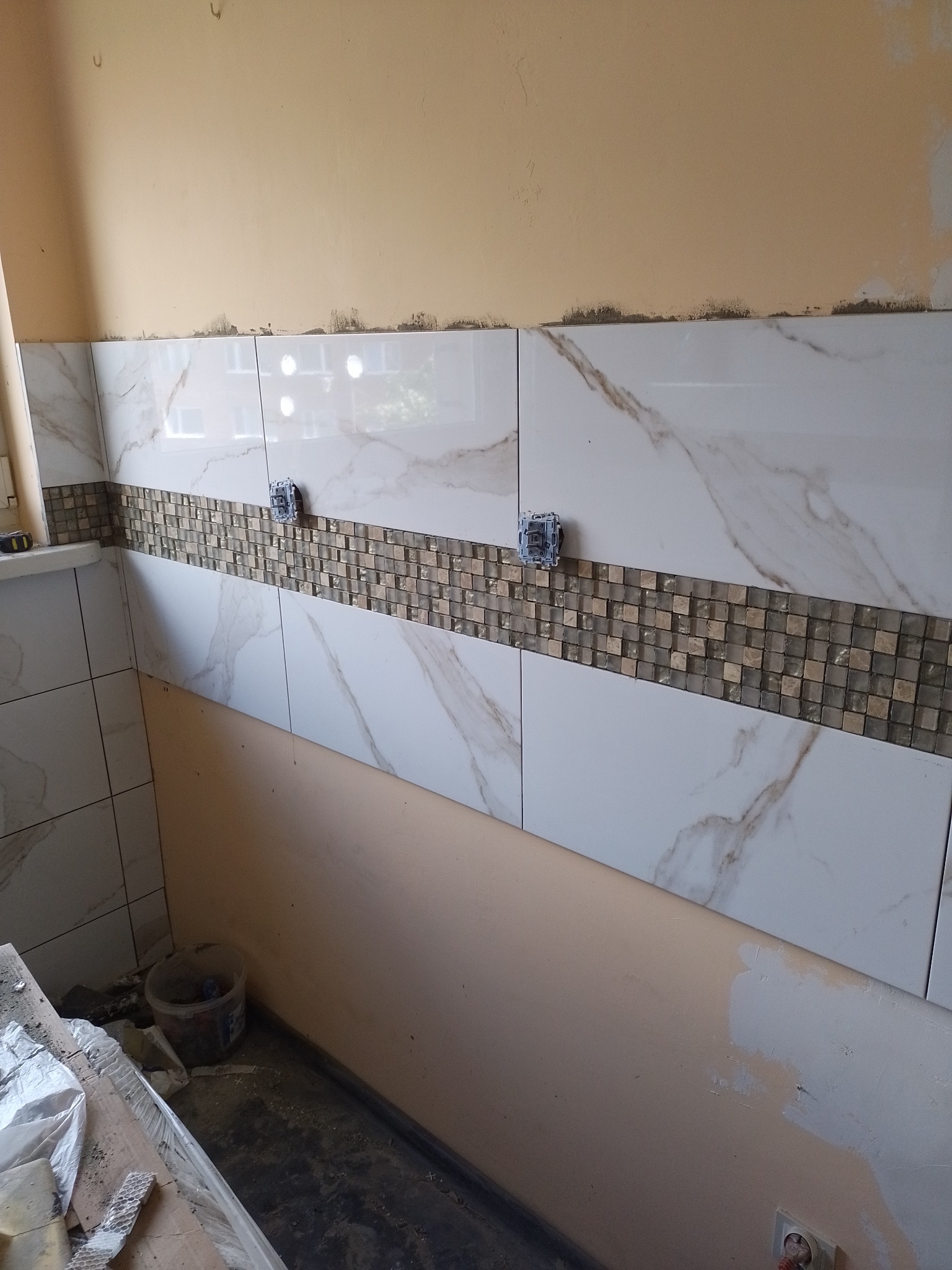 Fragment ściany w trakcie kładzenia płytek w kuchni lub łazience, widoczne białe płytki imitujące marmur i dekoracyjny pas mozaiki, puszki elektryczne wystające ze ściany, ślady kleju...