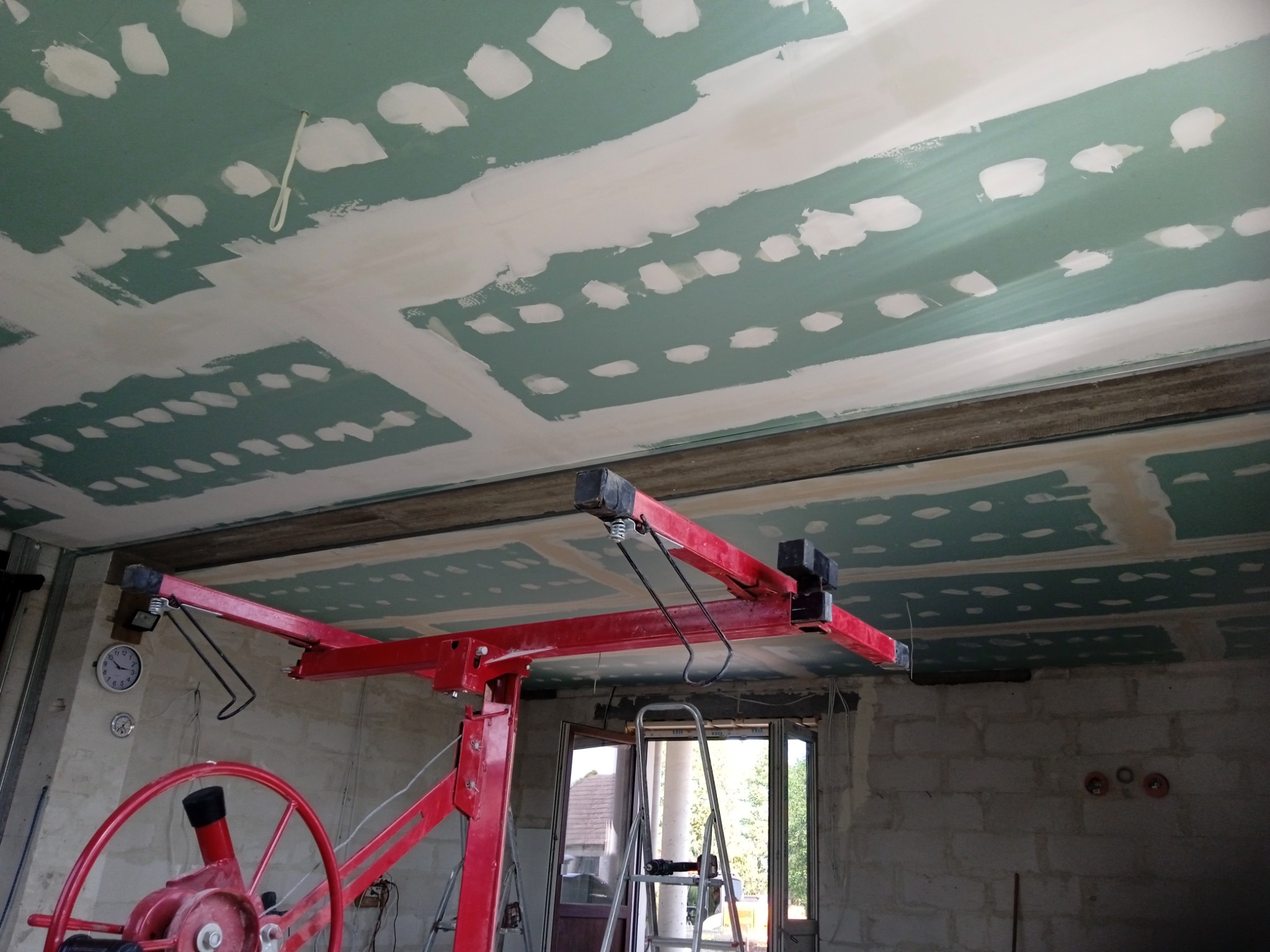 Montaż płyt kartonowo-gipsowych na suficie przy użyciu windy do płyt, widoczne szpachlowane łączenia i fragment ściany z surowego betonu komórkowego