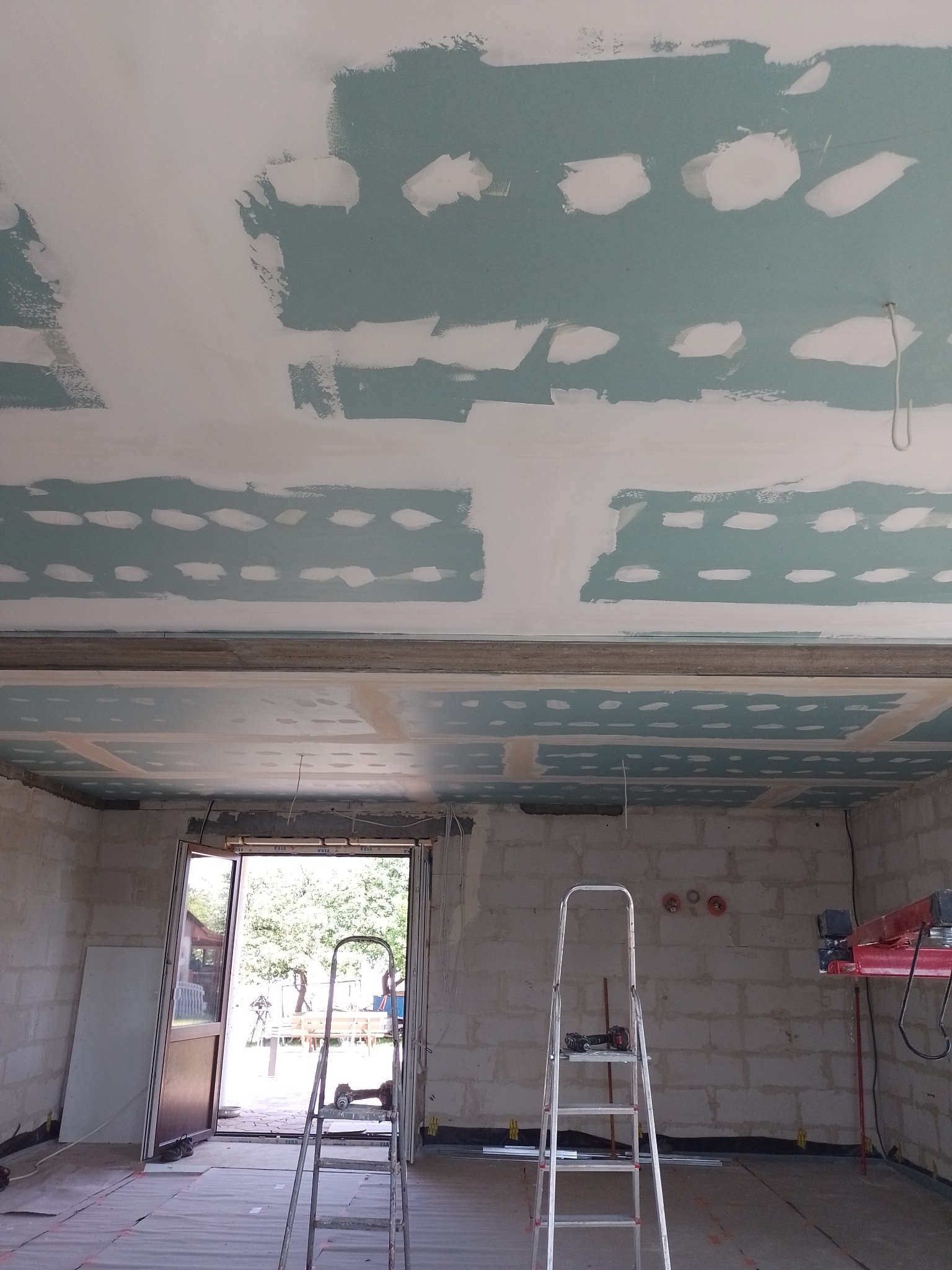 Sufit z płyt kartonowo-gipsowych w trakcie szpachlowania, widoczne łączenia płyt pokryte gładzią, pomieszczenie w trakcie remontu, drabiny w tle.