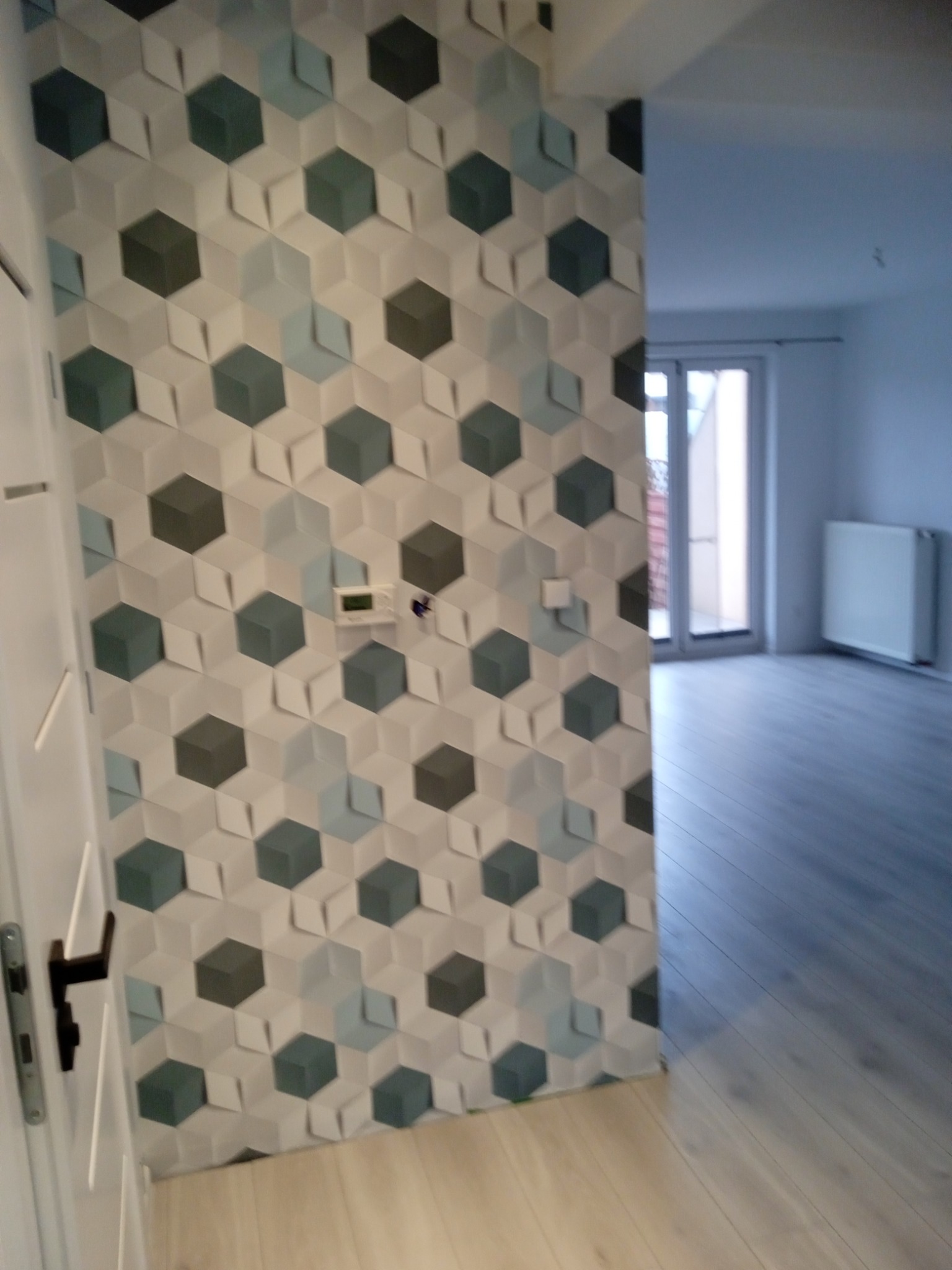 Ściana wykończona dekoracyjną tapetą w geometryczne wzory, widoczny fragment drzwi z klamką oraz fragment podłogi z panelami, w tle jasne pomieszczenie z oknem i grzejnikiem.