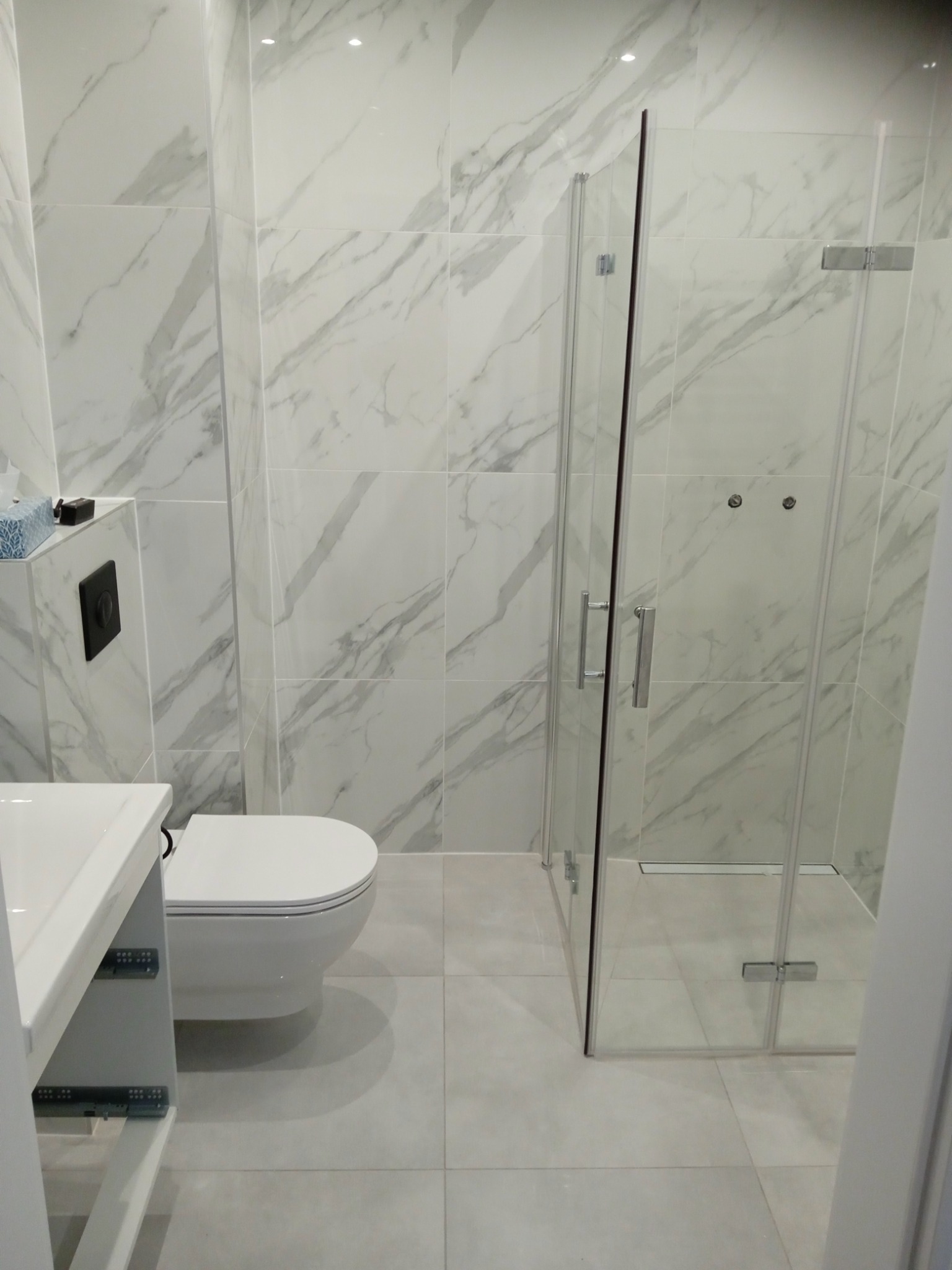 Nowoczesna łazienka z kabiną prysznicową ze szkła, umywalką z szafką i podwieszaną toaletą na tle płytek imitujących marmur.