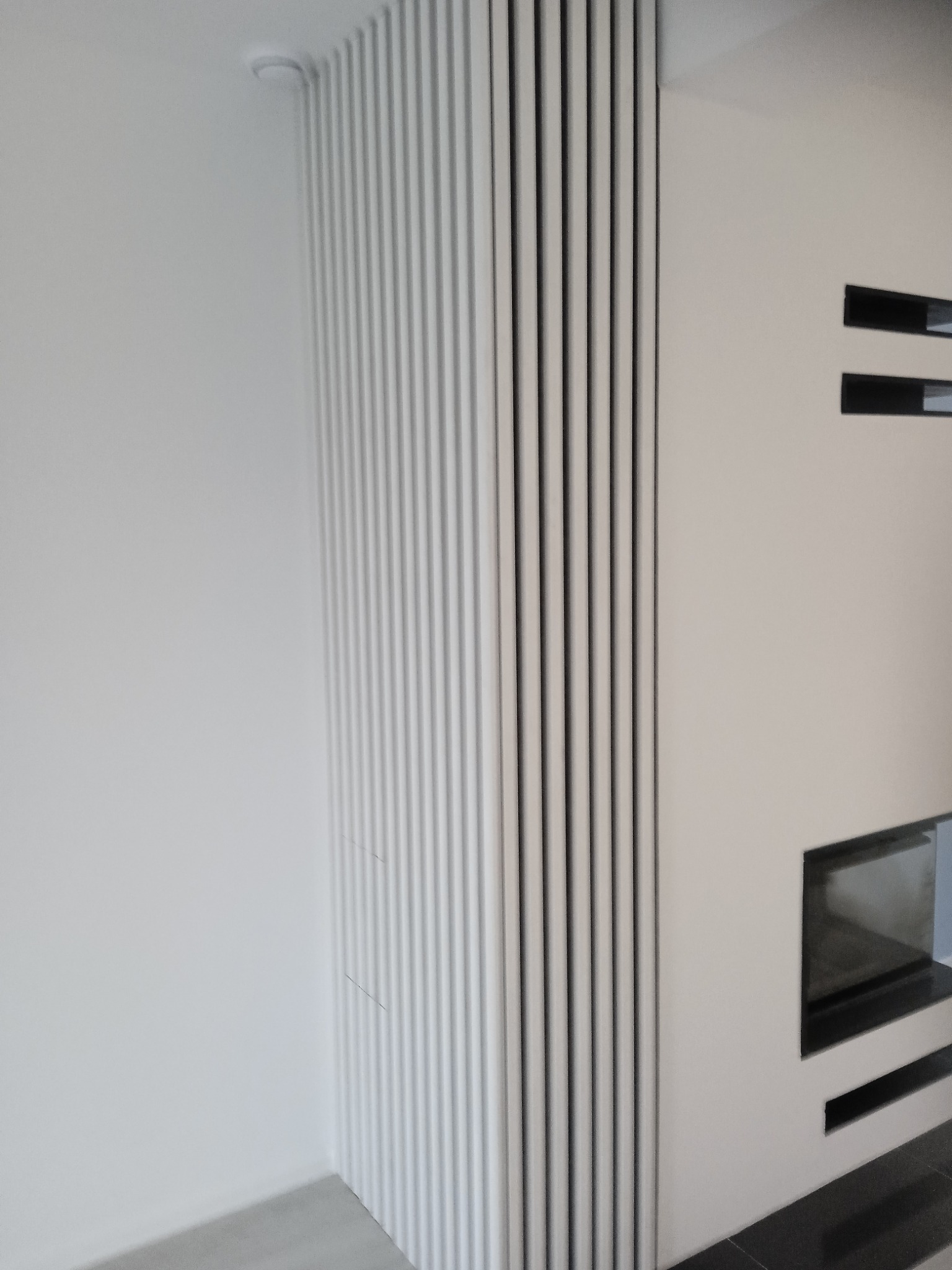 Nowoczesny salon z dekoracyjną ścianą z białych, pionowych paneli, minimalistyczny kominek z czarną obudową i wnękami, jasne ściany.