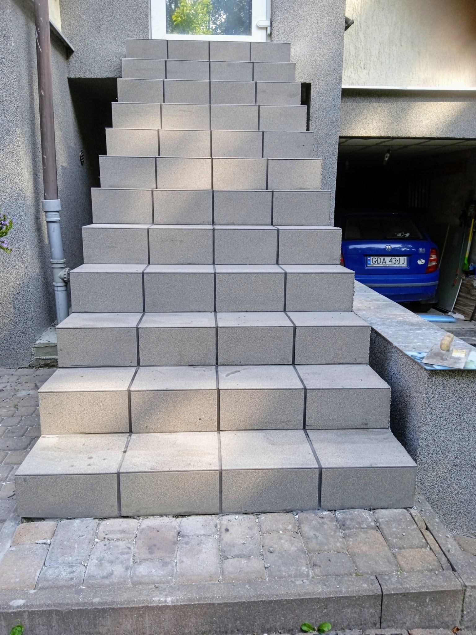 Zewnętrzne schody wejściowe wyłożone szarą, kwadratową płytką, prowadzące do drzwi i garażu z zaparkowanym niebieskim samochodem.