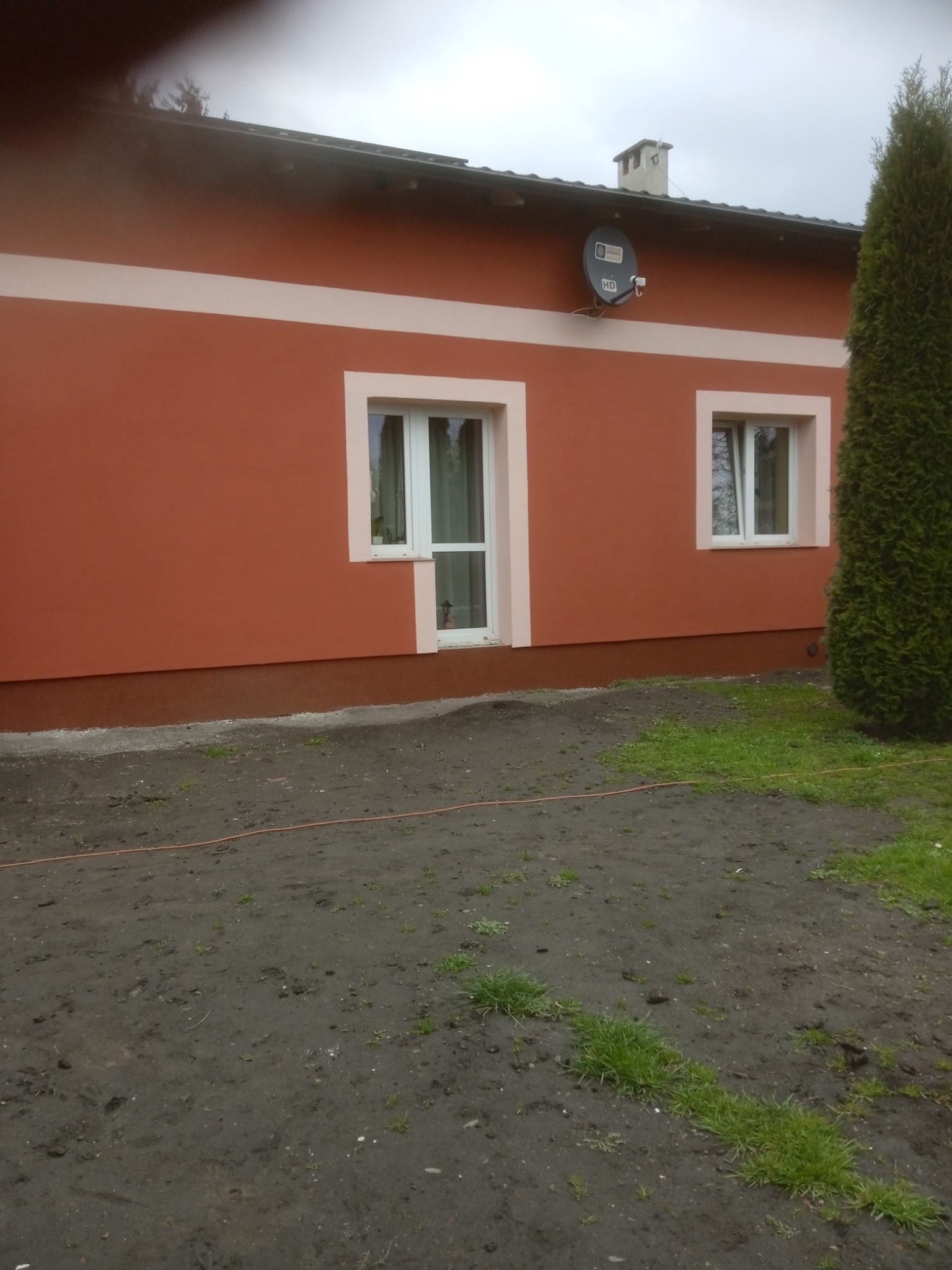 Ceglastoczerwona elewacja domu jednorodzinnego z białymi obramowaniami okien i poziomym pasem, widoczna antena satelitarna, brudne podwórko.