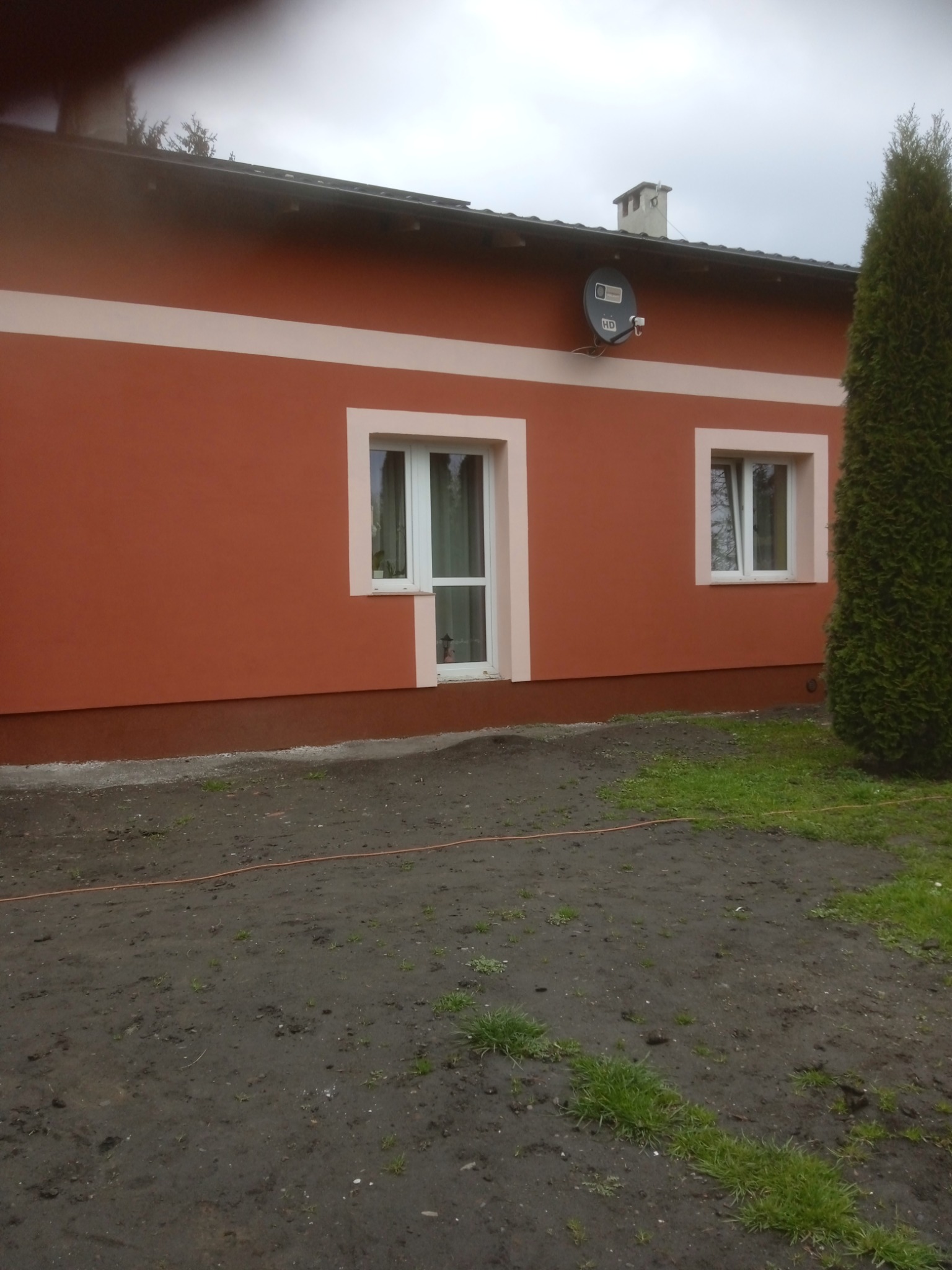 Ocieplony dom z pomarańczową elewacją, białymi obramowaniami okien, brązowym cokołem i ozdobnym pasem. Widoczna antena satelitarna na ścianie i tuja obok budynku.