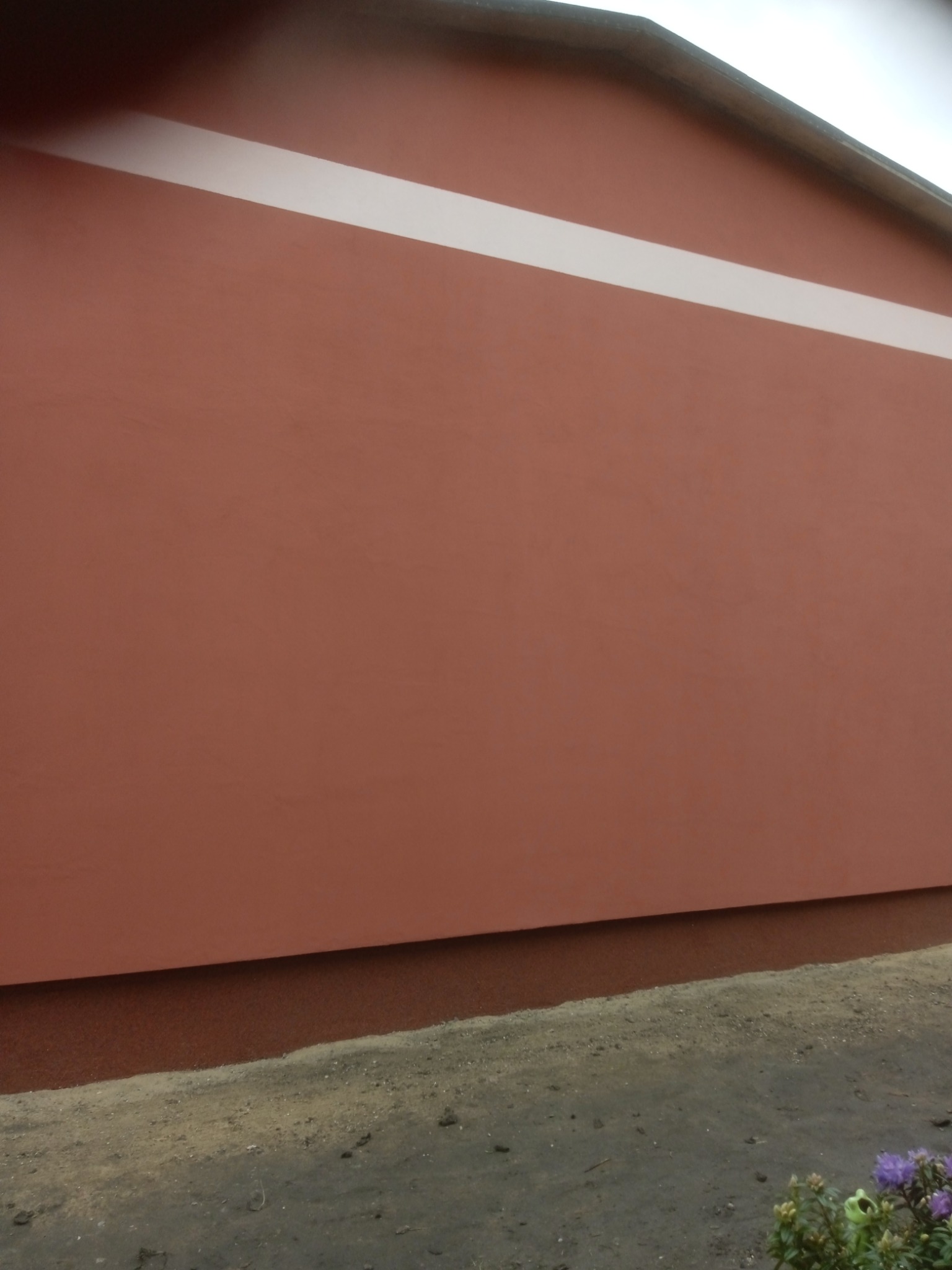 Pomarańczowa elewacja budynku z białym pasem dekoracyjnym, widoczne kwiaty przy fundamencie.