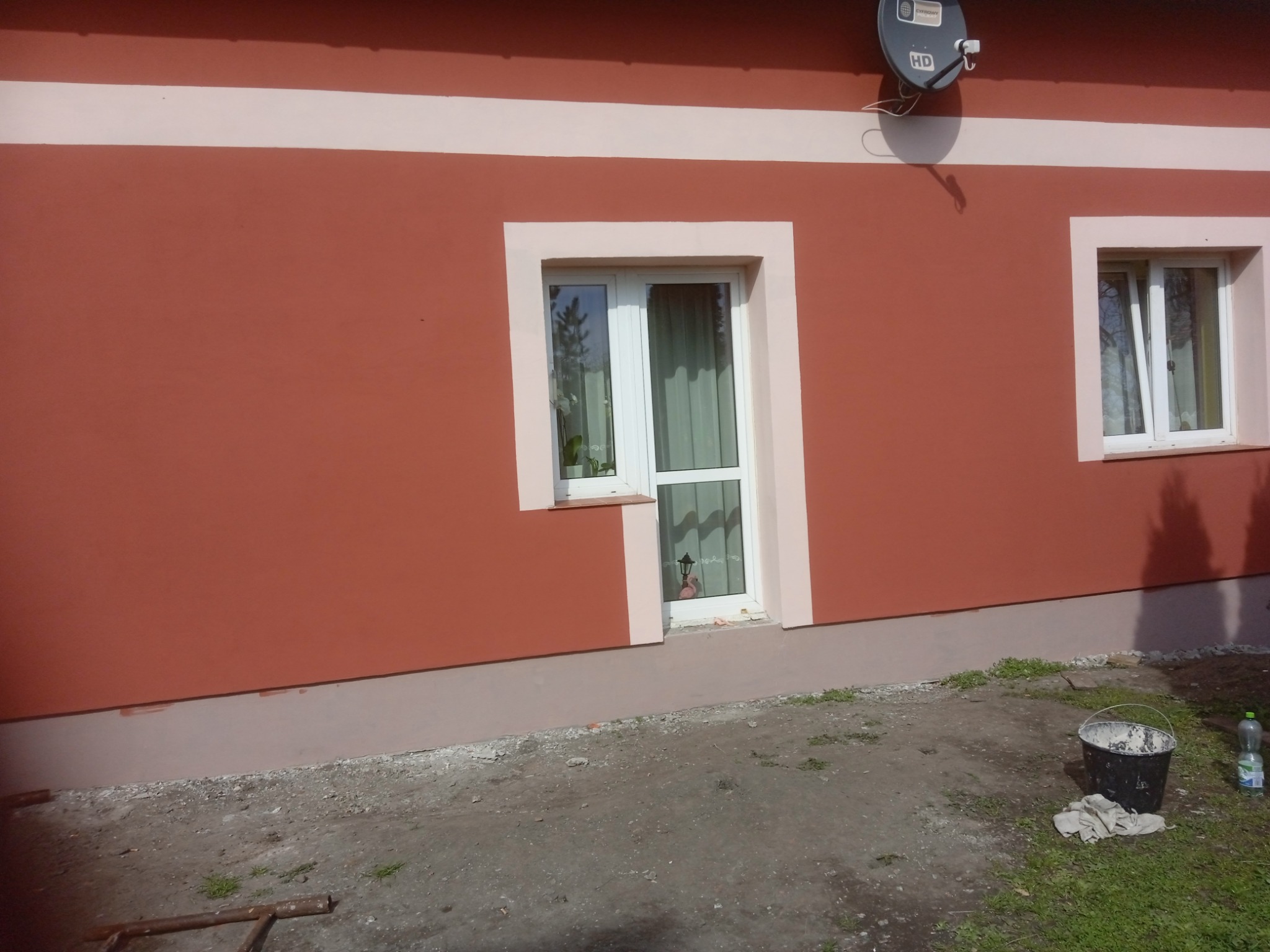 Ściana domu w Gdańsku po malowaniu elewacji na kolor ceglasty z białymi obramowaniami okien i pasem pod dachem, widoczny fragment terenu z wiadrem i butelką.