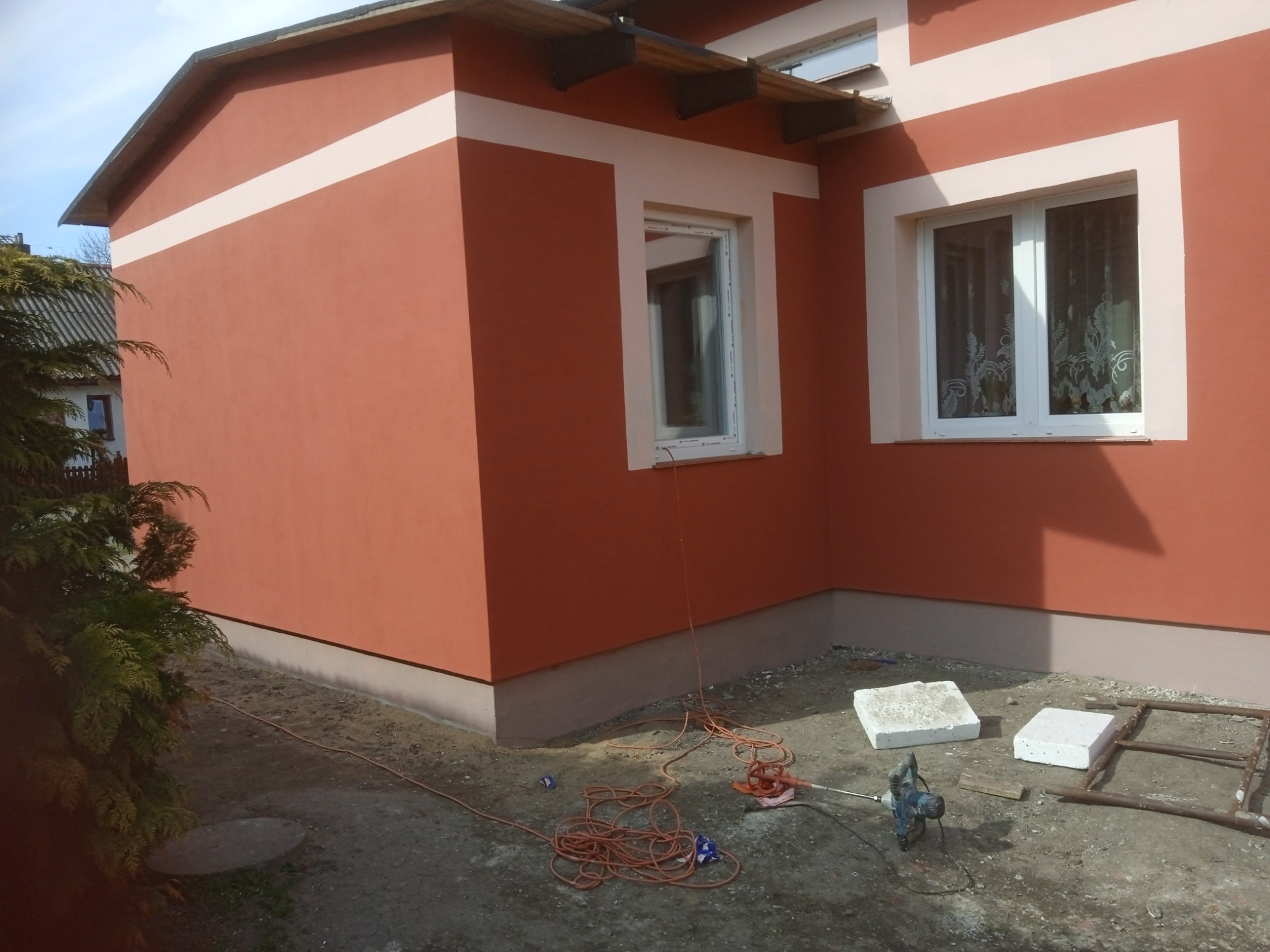 Świeżo pomalowana elewacja domu w odcieniach terakoty z kremowymi akcentami wokół okien i na pasie pod dachem, widoczne narzędzia i materiały budowlane na ziemi sugerują prace w toku.