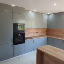 Nowoczesna kuchnia w odcieniach szarości z drewnianymi akcentami na blacie i panelu ściennym, oświetlenie punktowe w suficie i wyspa kuchenna z blatem drewnopodobnym na pierwszym planie.