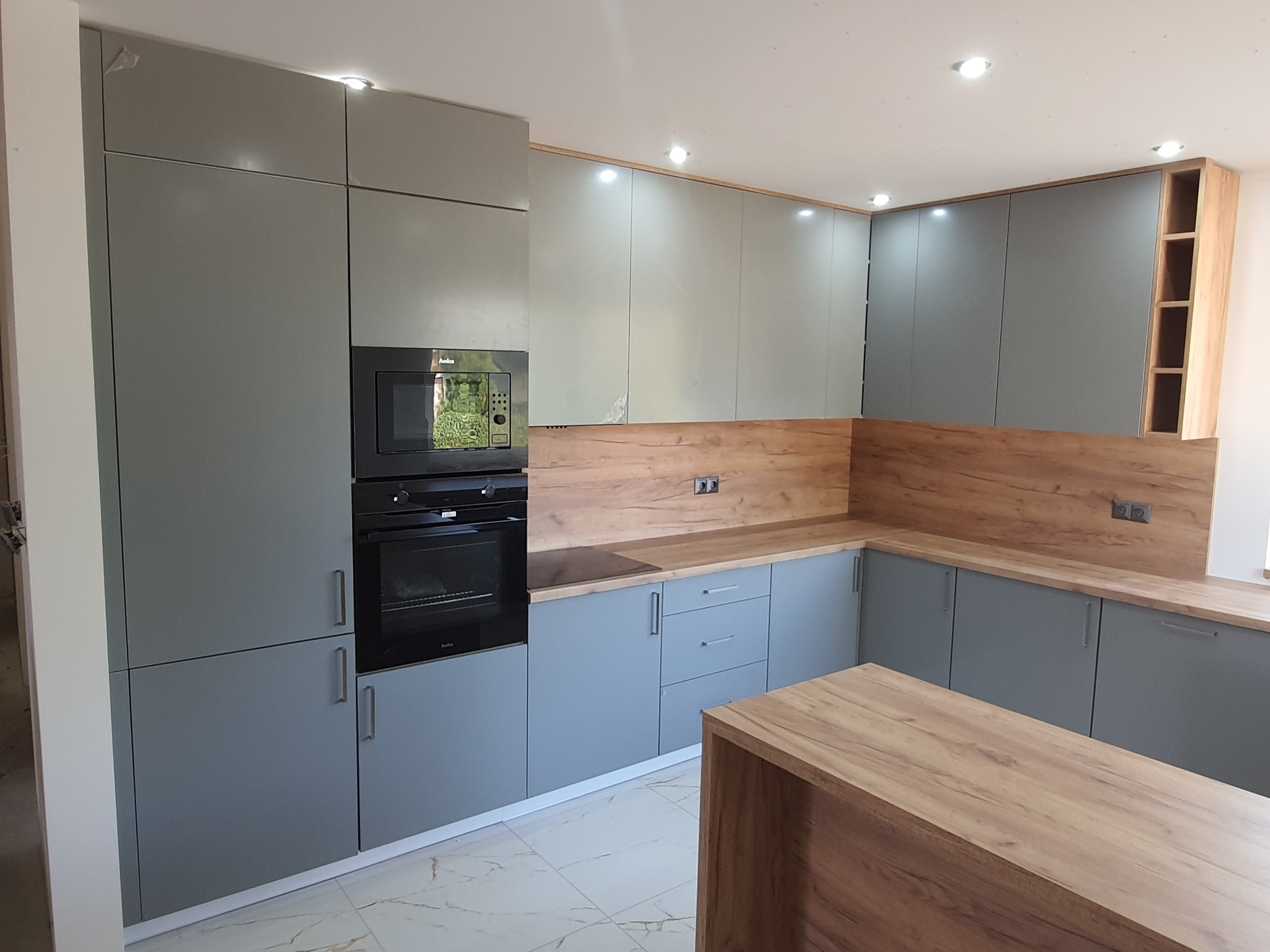 Nowoczesna kuchnia w odcieniach szarości z drewnianymi akcentami na blacie i panelu ściennym, oświetlenie punktowe w suficie i wyspa kuchenna z blatem drewnopodobnym na pierwszym planie.