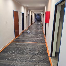 FIRMA FENSTER UND BAU Ewa Siegler - Korytarz w trakcie przygotowań do malowania: podłoga zabezpieczona folią, listwy przypodłogowe i framugi oklejone taśmą malarską, wiadro z farbą na środku, ściany przygotowane do malowania.