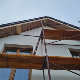 FIRMA FENSTER UND BAU Ewa Siegler - Fragment elewacji domu z widocznymi drewnianymi oknami, rusztowaniem i podbitką dachową w trakcie montażu.