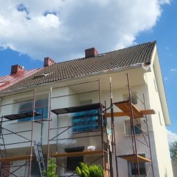FIRMA FENSTER UND BAU Ewa Siegler - Dwukondygnacyjny dom w trakcie malowania elewacji, z widocznymi rusztowaniami, zabezpieczonymi oknami folią i częściowo pomalowaną fasadą na jasny kolor, kontrastującą z czerwoną częścią dachu...