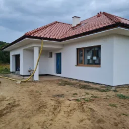 Nowoczesny dom jednorodzinny z białą elewacją i czerwoną dachówką, widoczny żółty wąż ogrodowy, teren wokół domu w trakcie prac wykończeniowych.