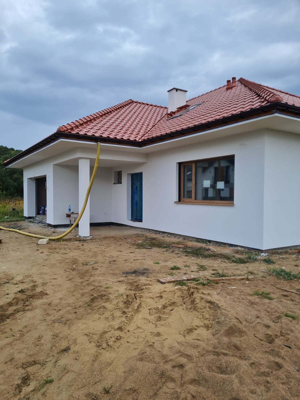 Nowo wybudowany dom jednorodzinny z białą elewacją i czerwoną dachówką ceramiczną, widoczne okno z drewnianą ramą i niebieskie drzwi wejściowe zabezpieczone folią, teren wokół domu piaszczysty,...