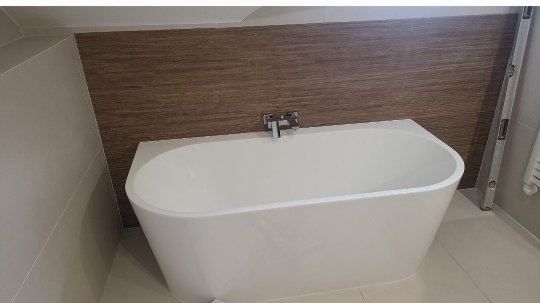 Biała, wolnostojąca wanna w łazience z brązową, drewnopodobną okładziną ścienną i poziomnicą opartą o ścianę. Nowoczesna armatura. Widoczne płytki podłogowe w kolorze beżowym.