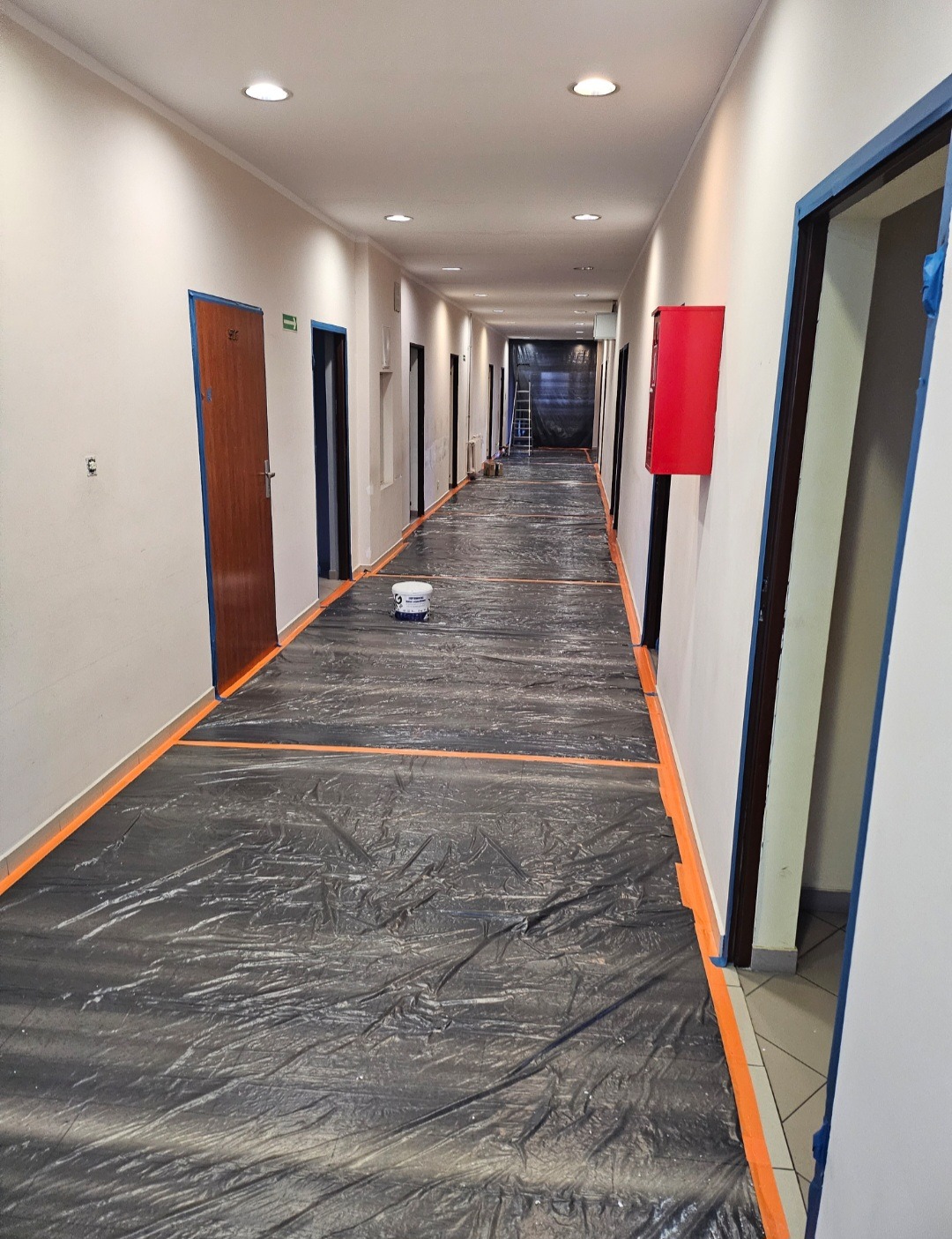 Korytarz w trakcie przygotowań do malowania: podłoga zabezpieczona folią, listwy przypodłogowe i framugi oklejone taśmą malarską, wiadro z farbą na środku, ściany przygotowane do malowania.