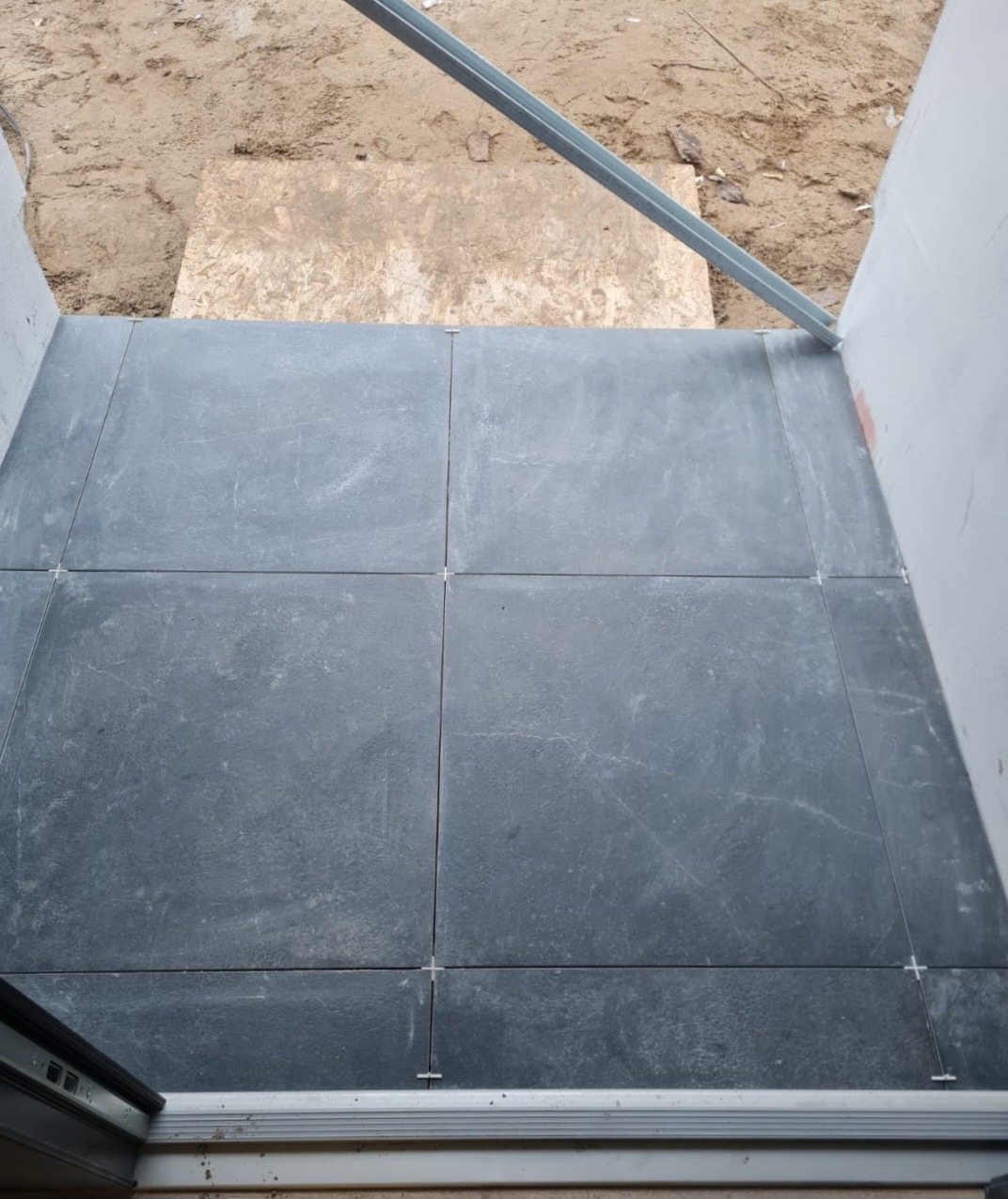 Ułożone, ciemnoszare płytki podłogowe z krzyżykami dystansowymi, widoczne elementy konstrukcyjne i niezagospodarowane podłoże.