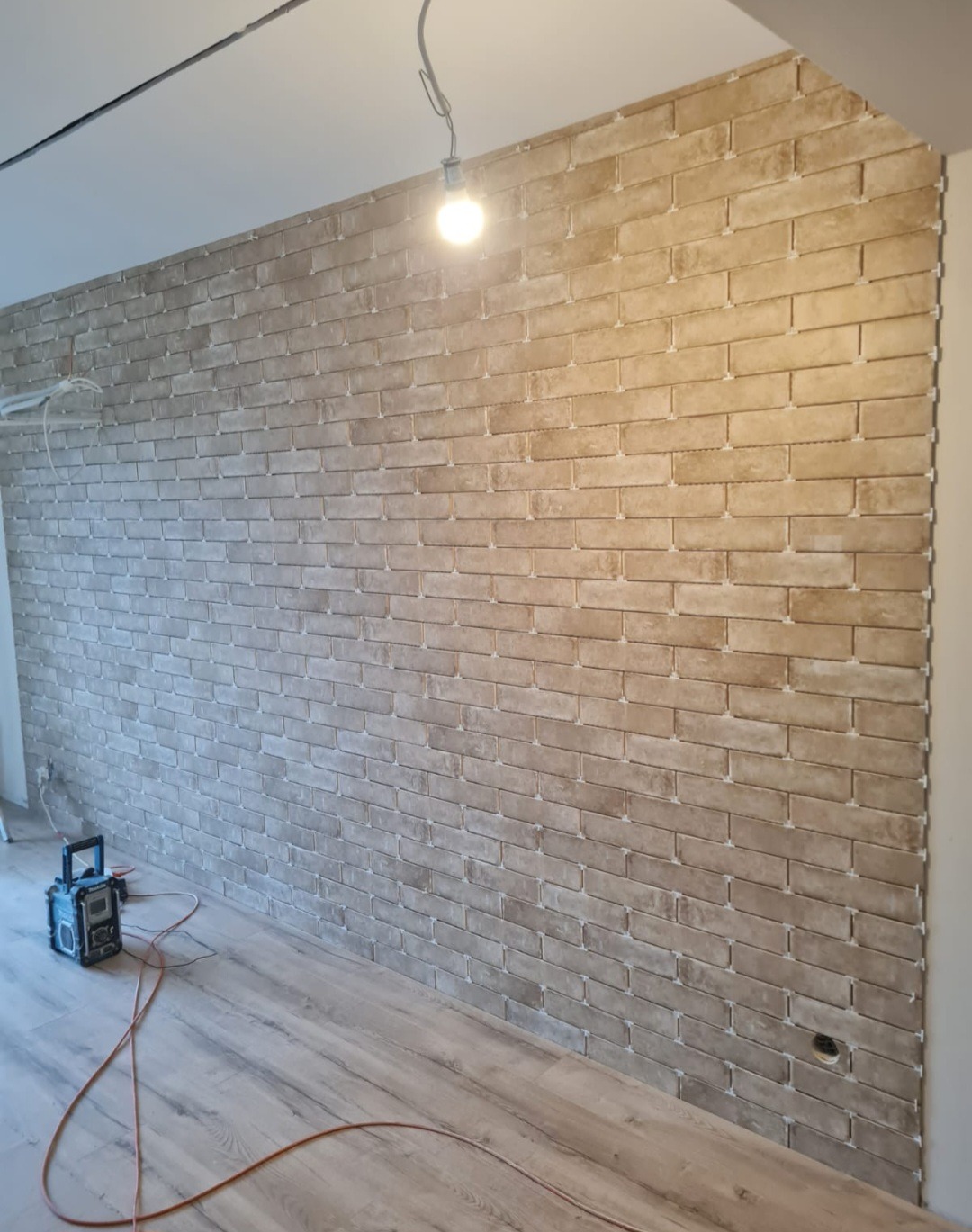 Ściana wyłożona beżową cegłą klinkierową, widoczne krzyżyki dystansowe, podłoga z paneli imitujących drewno, kabel elektryczny na podłodze i lampa wisząca z żarówką