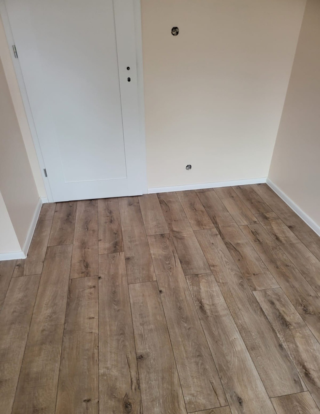 Nowo wykończone wnętrze z panelami podłogowymi imitującymi drewno, białe ściany z widocznymi puszkami instalacyjnymi i białe drzwi.