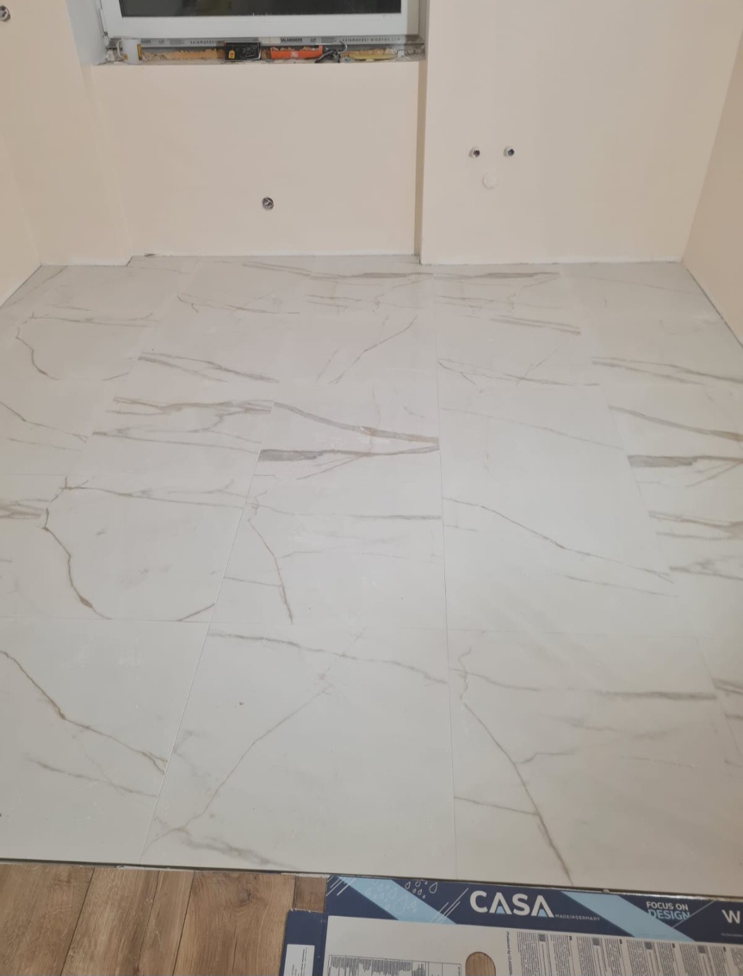 Ułożone białe płytki podłogowe z marmurkowym wzorem w jasnym pomieszczeniu, widoczne puszki elektryczne w ścianie i poziomica w oknie, fragment opakowania po płytkach w rogu.