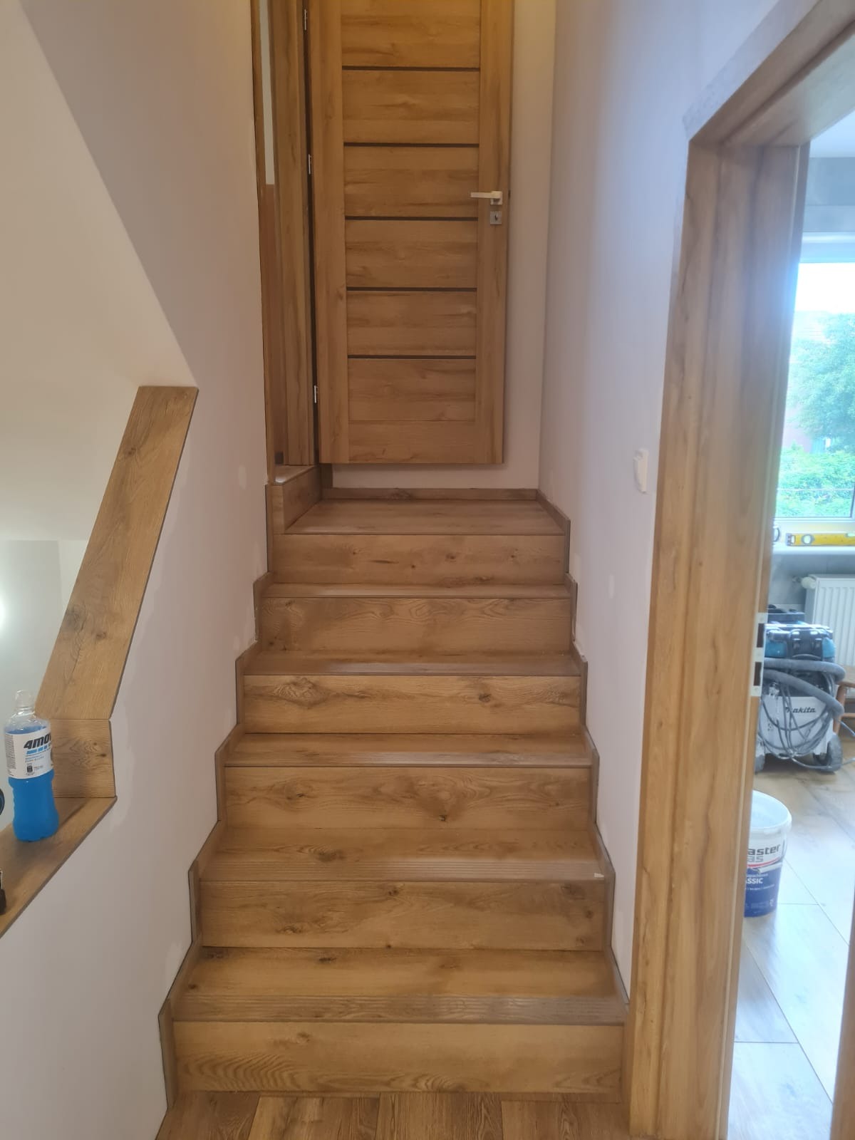 Drewniane schody z drzwiami w mieszkaniu, widoczne narzędzia i materiały malarskie w tle.