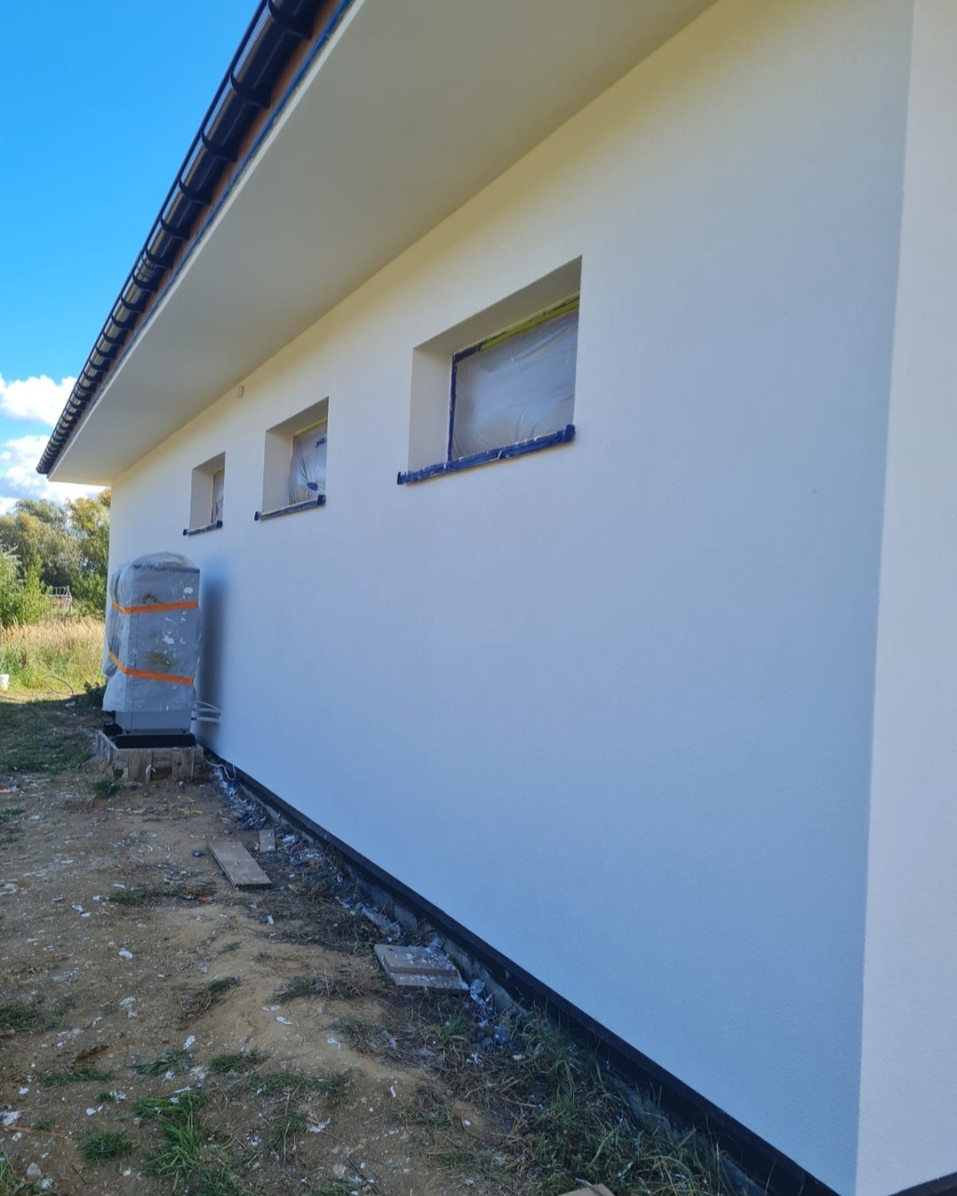 Świeżo pomalowana, biała elewacja budynku z zabezpieczonymi oknami folią malarską, widoczny klimatyzator owinięty folią ochronną.