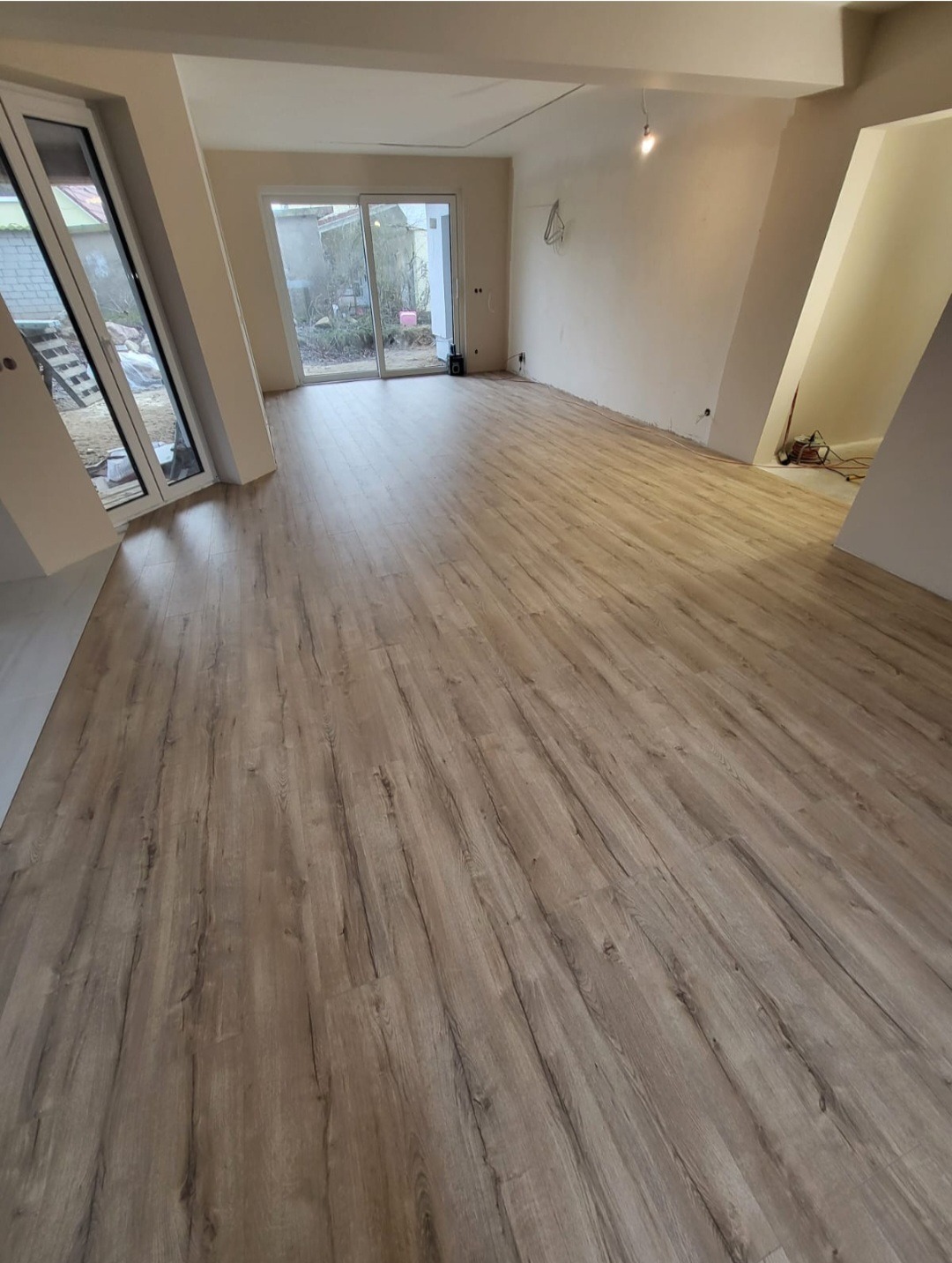 Wnętrze po remoncie z nową podłogą z paneli imitujących drewno, widoczne białe ściany, okna PCV i drzwi balkonowe, oraz niezabudowana wnęka.