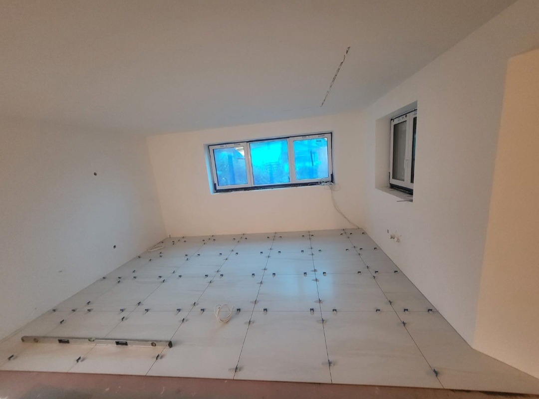 Pokój na poddaszu w trakcie układania płytek podłogowych z użyciem krzyżyków dystansowych, widoczna poziomica przy ścianie, okno PCV z niebieską folią ochronną.