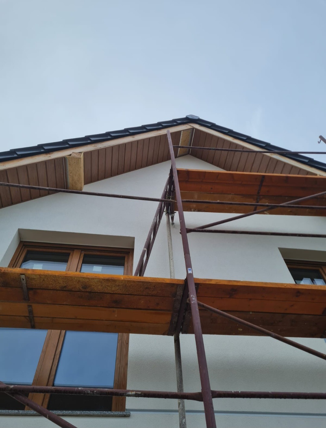 Fragment elewacji domu z widocznymi drewnianymi oknami, rusztowaniem i podbitką dachową w trakcie montażu.
