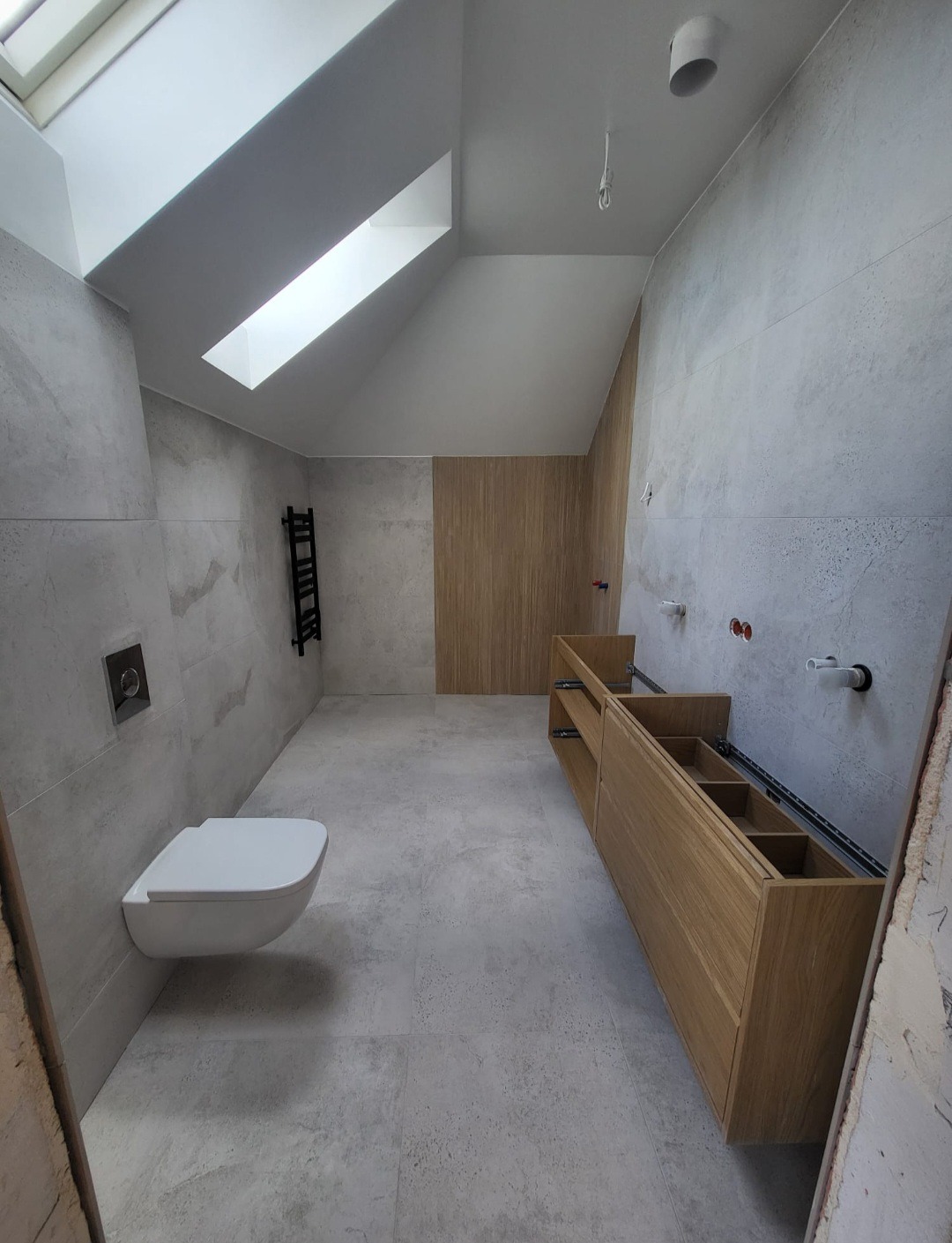 Nowoczesna łazienka w trakcie wykańczania: podwieszana toaleta, drewniana szafka pod umywalkę z otwartymi półkami, szare płytki imitujące beton, grzejnik drabinkowy i okno dachowe.