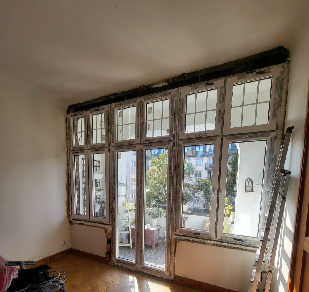 Montaż białych okien PCV z widokiem na balkon i budynki w tle, widoczna drabina oparta o ścianę, podłoga parkiet.