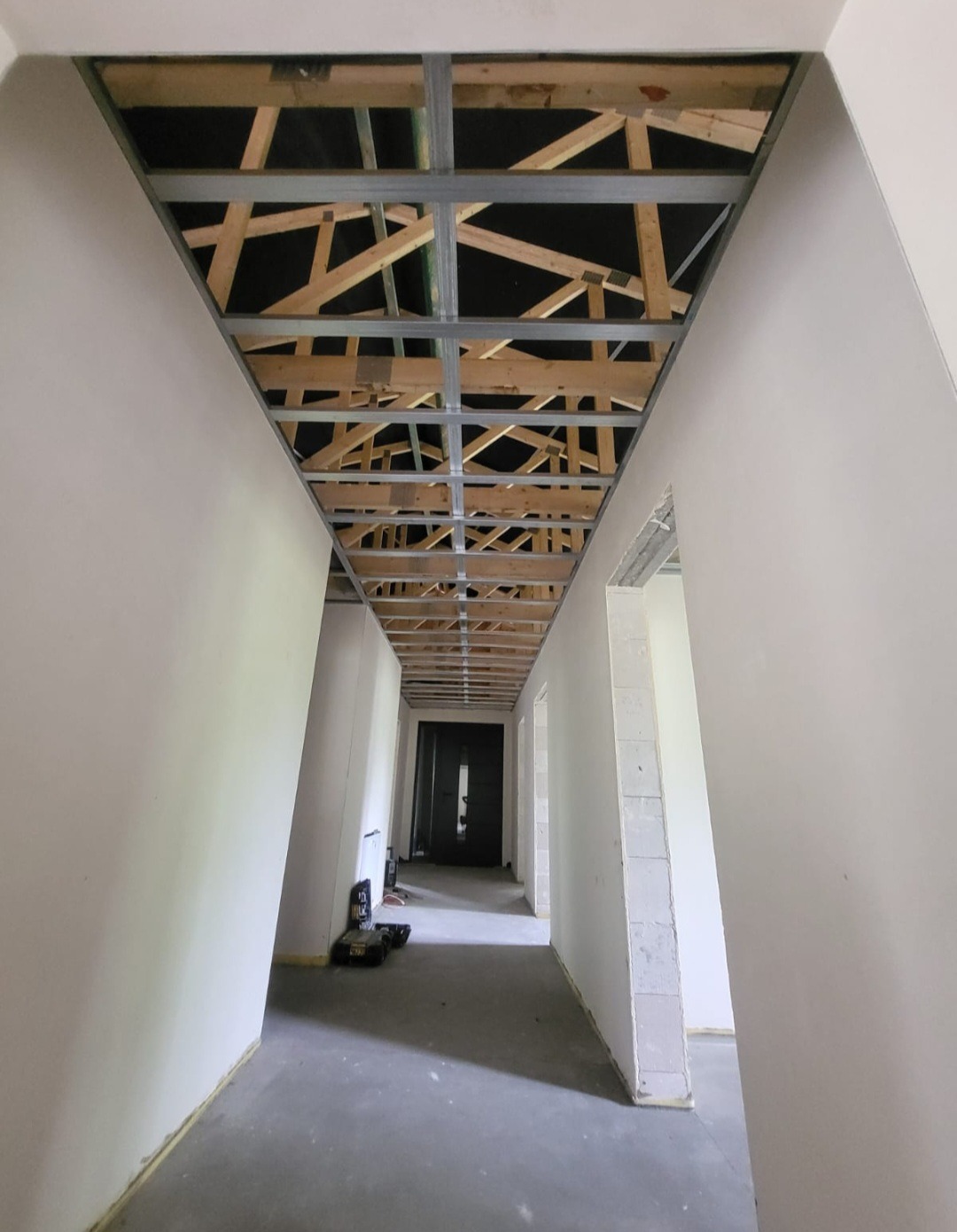 Korytarz w trakcie prac wykończeniowych: widoczne metalowe profile sufitowe na drewnianej konstrukcji, surowe ściany w kolorze białym, betonowa podłoga, narzędzia na podłodze w oddali.