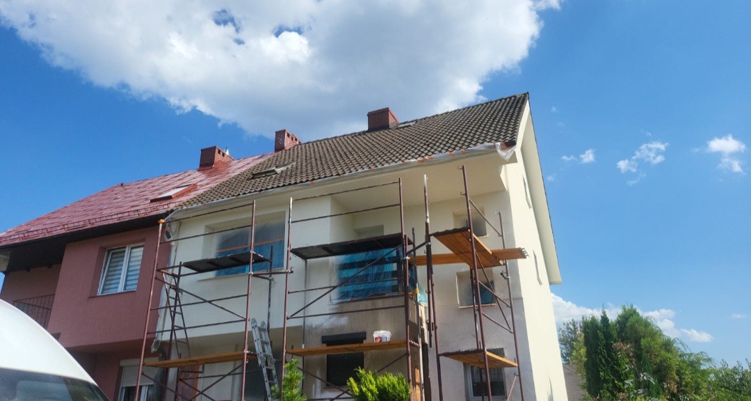 Dwukondygnacyjny dom w trakcie malowania elewacji, z widocznymi rusztowaniami, zabezpieczonymi oknami folią i częściowo pomalowaną fasadą na jasny kolor, kontrastującą z czerwoną częścią dachu...