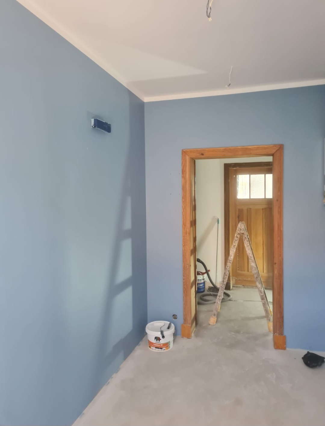 Świeżo pomalowane na niebiesko ściany w mieszkaniu w Dobrej, z widoczną drewnianą futryną drzwi, drabiną i wiadrem z farbą w rogu.