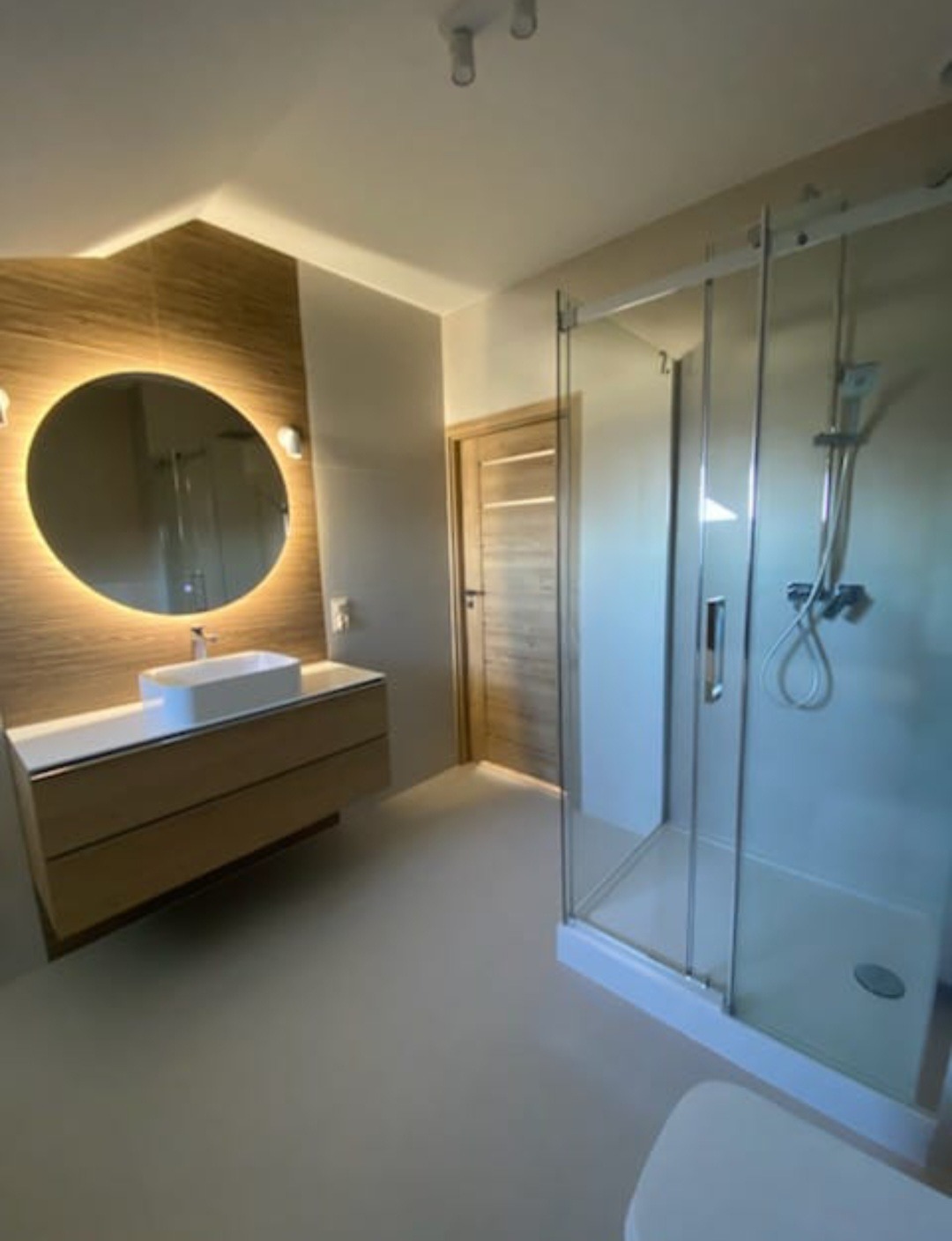 Nowoczesna łazienka z podświetlanym lustrem, drewnianą szafką pod umywalką i kabiną prysznicową ze szklanymi drzwiami przesuwnymi.